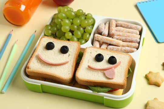 Sandwiches with smiley faces in lunch box, bottle of juice and stationery on yellow table, closeup