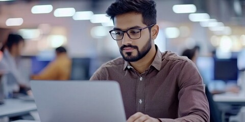 Fototapeta premium Portrait of a young male programmer in a brown shirt and glasses who is concentrating on working on a laptop while sitting at a desk in the office， Work Culture, and Professional Dedication for Labor 