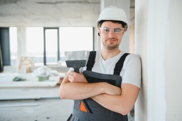 Worker in uniform plastering wall with putty knife indoors