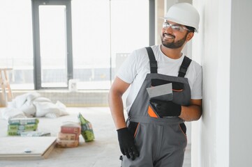 Indian Male builder in work overalls plastering a wall using a construction trowel.
