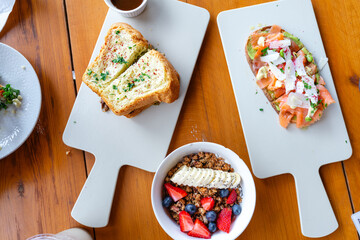 Brotzeit in einem Restaurant oder Kaffee ein Brot mit Lachs, ein Sandwich zusammen gefaltet, dekoriert mit grünen Kräutern und eine ACAI Bowl mit frischen Beeren und Banane auf Holztisch