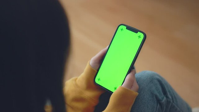 Hand of woman holding mobile smartphone with blank green screen mock up while sitting on a sofa at home