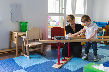 Young cute boy with cerebral palsy practices standing and balancing using handrails with assistance...