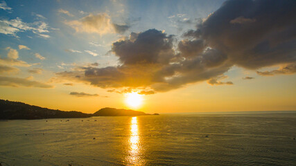 A breathtaking aerial view of a coastal bay at sunset, with golden light reflecting on the calm sea and silhouetted hills in the distance. beautiful reflection of the golden sun at Patong Bay Phuket.