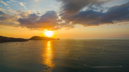 A breathtaking aerial view of a coastal bay at sunset, with golden light reflecting on the calm sea and silhouetted hills in the distance. beautiful reflection of the golden sun at Patong Bay Phuket.