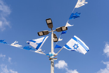Israeli flags and blue-and-white decorations adorn a streetlight pole against a bright blue sky....