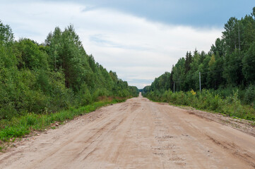 Long straight country dirt road across the forest. Green trees along the roadside, cloudy sky, summertime