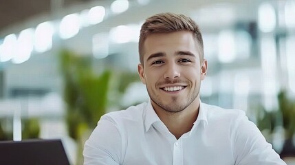 Fototapeta premium A young businessman sits at a sleek office desk, working on a laptop and smiling joyfully at the camera. He is focused on digital marketing and design concepts in a contemporary environment