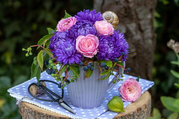 Blumen-Arrangement mit pink Rosen, lila Sommerastern und Wildem Wein in einer Tasse im Garten