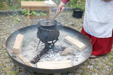 Hand stirring a black cauldron over a fire pit, creating an authentic medieval cooking experience. The scene is set on cobblestones, surrounded by wood logs, evoking historical traditions