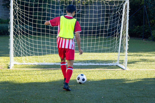 Playing soccer, boy in red uniform kicking ball towards goal on field