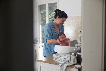 Mother bathing newborn in baby bathtub in kitchen, indoor scene, baby looking calm, maternal care, nurturing moment, home environment, hygiene routine, parental bonding, infant bathing