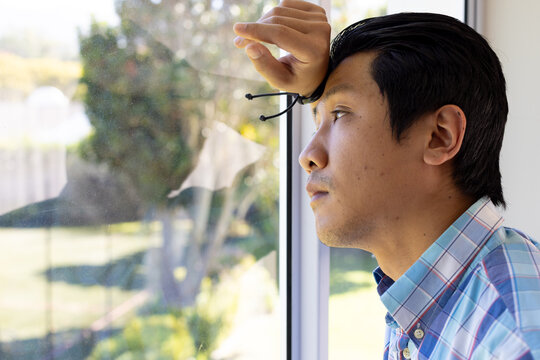 Looking out window, asian man in plaid shirt contemplating, resting head on hand, copy space