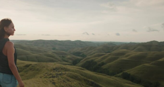 Young woman is embracing the beauty of nature while standing on a hilltop, gazing at the mesmerizing expanse of the wairinding hills in sumba island, indonesia