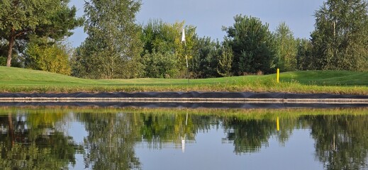 A golf green with a white flag near a pond, reflecting trees and the sky in the water. Peaceful scene with natural colors.