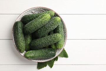 Fresh cucumbers in bowl and green leaves on white wooden table, top view. Space for text