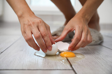 Woman removing shell and broken egg from grey wooden floor indoors, closeup