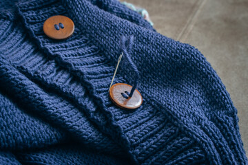Blue wool knitted cardigan with wooden button and sewing needle. Knitting rib pattern with buttons. Handmade cardigan. Warm clothes background. Autumn fashion. Needlework, close up.