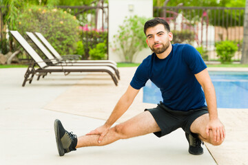 Active young man in workout attire stretching his legs poolside in the backyard of a home