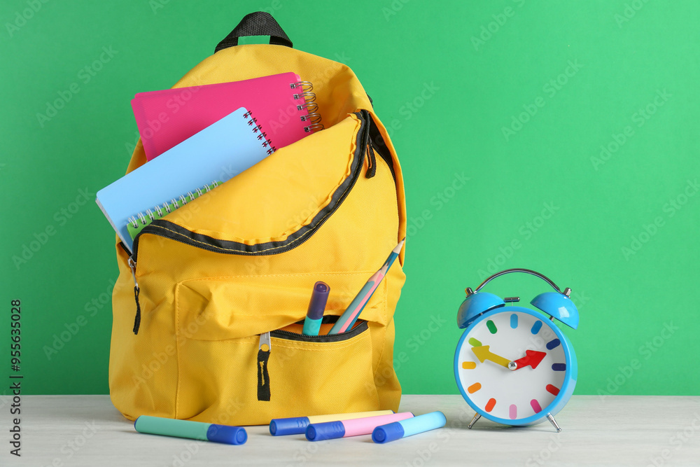 Poster backpack with different school stationery and alarm clock on white table against green background