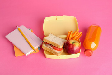 Lunch box with snacks, bottle of juice and stationery on pink background