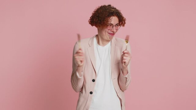 Ready to eat. Hungry Caucasian man with curly hair waiting for serving dinner dishes with with restlessness holding cutlery fork knife, will appreciate delicious restaurant meal on pink background