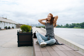 Relaxed redhead female finding serenity on river pier, savoring tranquil atmosphere and cloudy sky. Pretty smiling young woman appears at ease, taking respite from urban living outdoors.