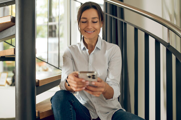 Smiling pretty woman sitting on a winding stairs with a phone in hand