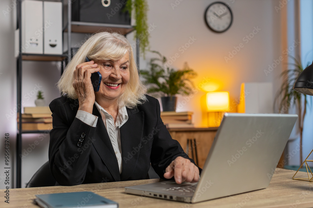 Wall mural Confident senior Caucasian businesswoman operator grandmother communicating with client by smartphone, making notes working on laptop computer at home office desk. Concept of distance job on Internet.