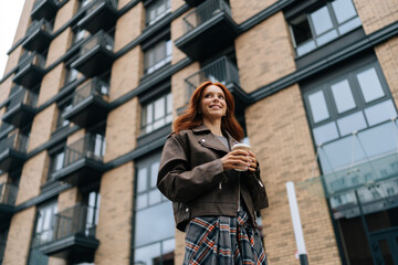 Fototapeta premium Low-angle view of cheerful redhead woman savoring coffee break in urban environment, holding cup and sipping drink with smile, exuding joy and relaxation, standing on background of modern building.