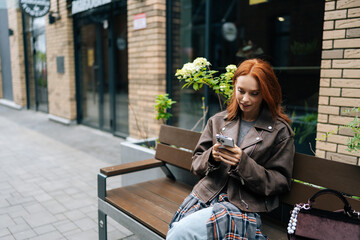 Pretty young woman in trendy leather jacket sitting on bench outdoors, happily browsing smartphone. Stylish redhead female using mobile phone on city bench, enjoying free time in European city.