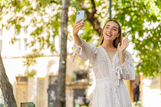Young pretty woman blogger taking selfie on smartphone, communicating video call online with subscribers, recording stories for social media vlog outdoors. Lovely girl standing in urban city street.