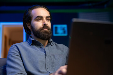 Man on couch working from home in apartment solving business related tasks on laptop. Teleworking company manager on couch in neon lit living room sending emails on notebook