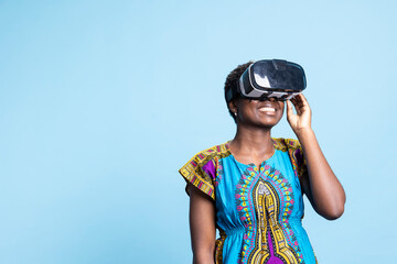 Young cheerful person enjoying artificial intelligence on vr glasses, using 3d interactive vision against blue background. Joyful girl having fun with virtual reality headset, futuristic tech.