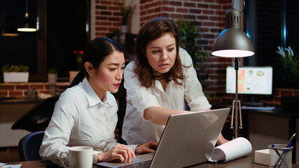 Asian businesswoman and colleague working together doing computer tasks for team project in office overnight. Coworkers looking over accounting figures on laptop screen late at night