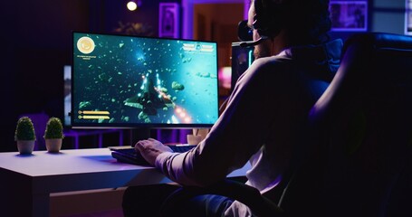 Man in dimly lit apartment playing singleplayer videogame with spaceship shooting laser streams at...