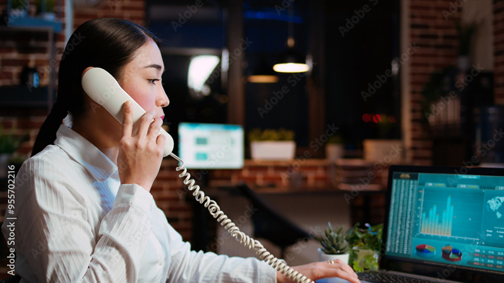 Aufkleber asian employee taking landline telephone call while working with financial statistics graphs on lapt