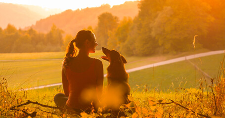 SILHOUETTE: Young woman and her dog look at each other while watching the sunset. Wonderful friendship and genuine connection between a lady and her furry pet in the embrace of beautiful autumn nature