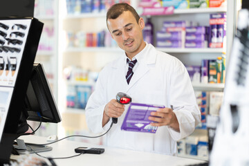 Smiling young man apothecary scanning bar-code standing at cash desk in chemist's shop