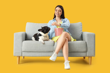 Young woman with her French bulldog eating popcorn on sofa against yellow background