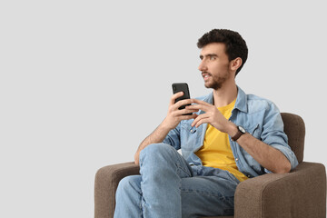 Handsome young man using mobile phone in armchair on light background