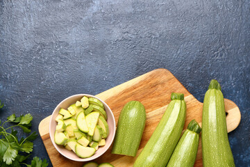 Board with fresh green zucchini and bowl of slices on blue background