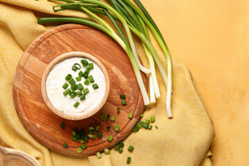 Board with bowl of tasty sour cream and sliced green onion on yellow background