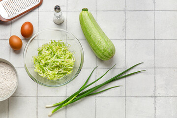 Bowl with grated zucchini and ingredients for preparing fritters on white tile background