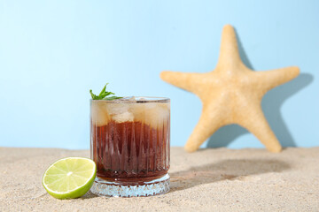 Glass of cold Cuba Libre cocktail and starfish on sand near blue background