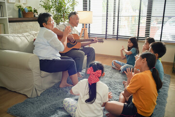 Elderly couple playing guitar and clapping along with a children seated on the floor in a cozy living room. The scene captures the joy of intergenerational bonding through music family atmosphere