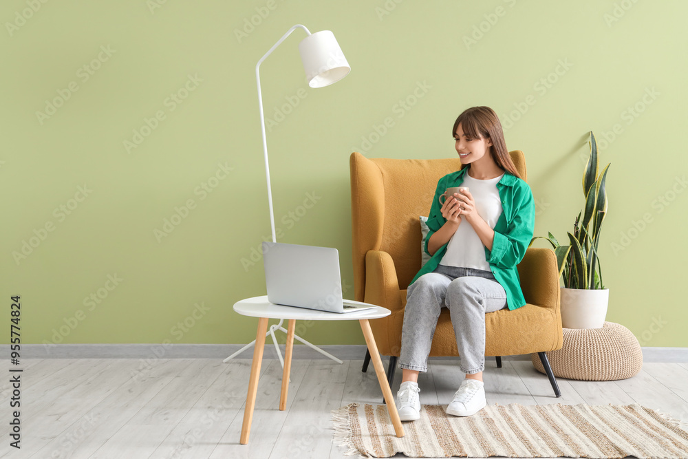 Poster Young woman with cup of coffee in armchair watching movie on laptop near green wall