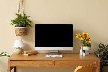 Beautiful workplace with modern computer, books and sunflowers near beige wall in room