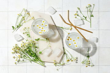 Composition with glasses of water, ice cubes and chamomile flowers on light tile background