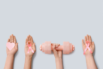 Female hands with pink ribbons, paper heart and dumbbell on grey background, closeup. Breast cancer awareness concept